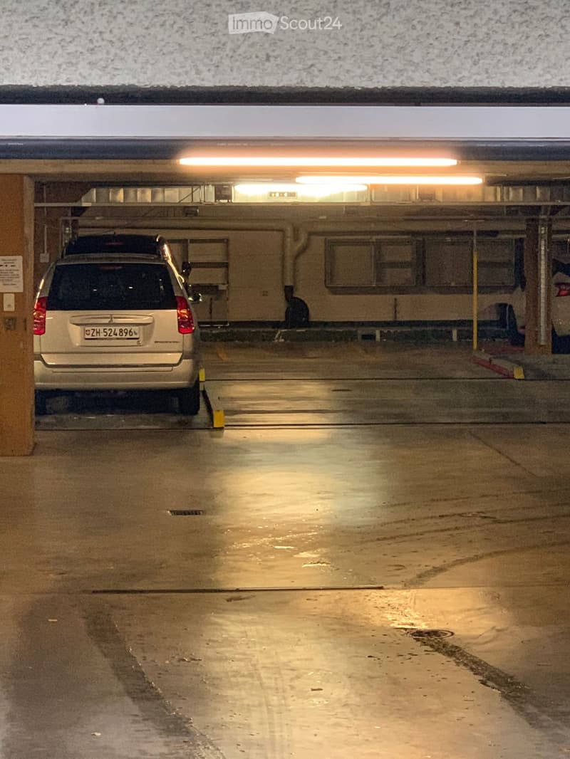 Underground parking area with a parked car, concrete pillars, and fluorescent lighting.