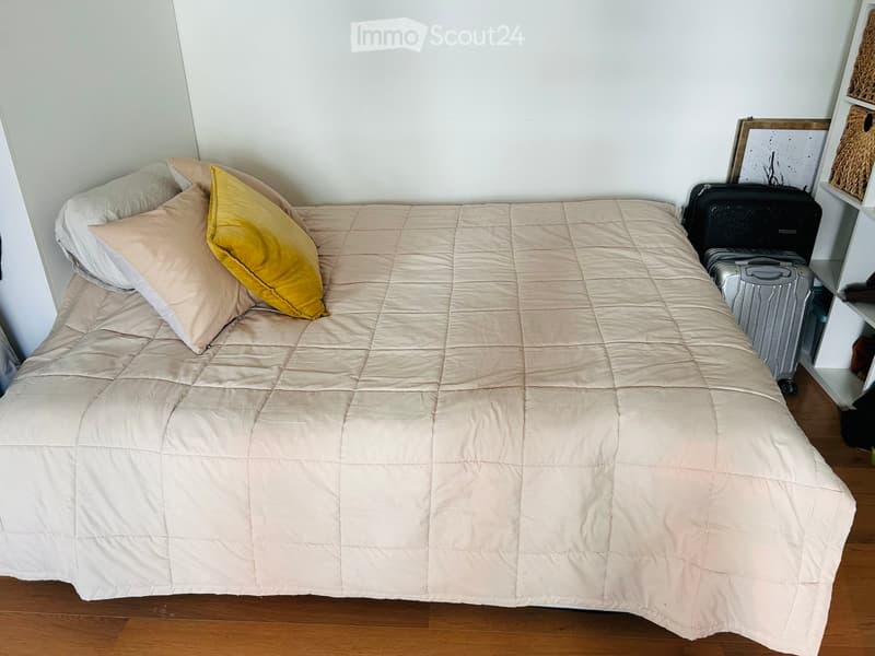 A neatly made bed with a beige duvet and pillows, including a yellow accent pillow, positioned against a white wall. A suitcase and a shelf with baskets are visible to the right.