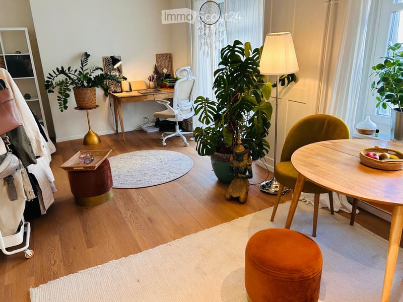 A cozy, well-lit room with wooden flooring, a round table with a green chair, a small desk with a chair, a large plant, and decorative items including a dreamcatcher and a small statue.