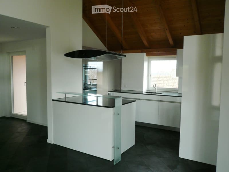 Modern kitchen with white cabinetry, a central island with a glass countertop, and a range hood above the stove area. The space features dark tiled flooring and large windows allowing natural light.