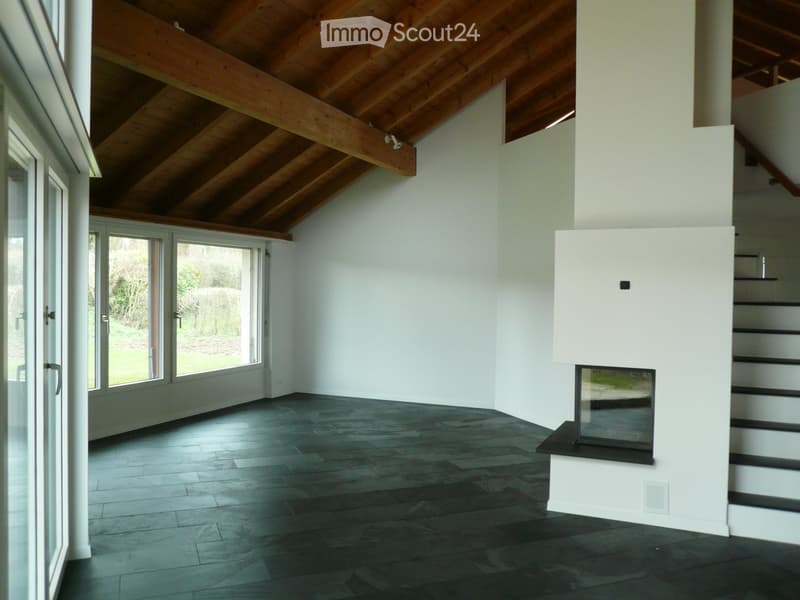 Bright, open living space with high wooden-beamed ceiling, dark hardwood floor, large glass doors leading to outdoor area, and a modern white fireplace with black surround.