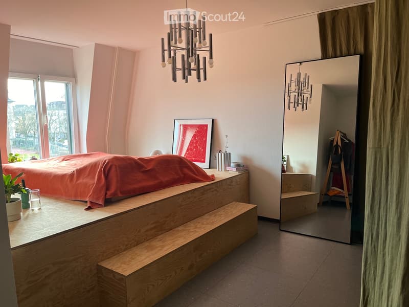 Modern bedroom with a raised wooden bed platform, large mirror, chandelier, and decorative items.