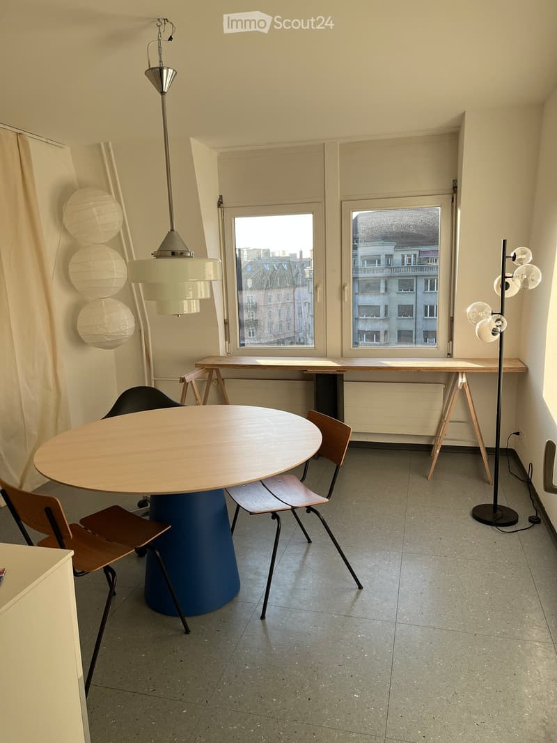 A modern, minimalist room with a round wooden table, four chairs, a tall floor lamp, and a window with a curtain. The room features a concrete floor and a view of a cityscape through the window.