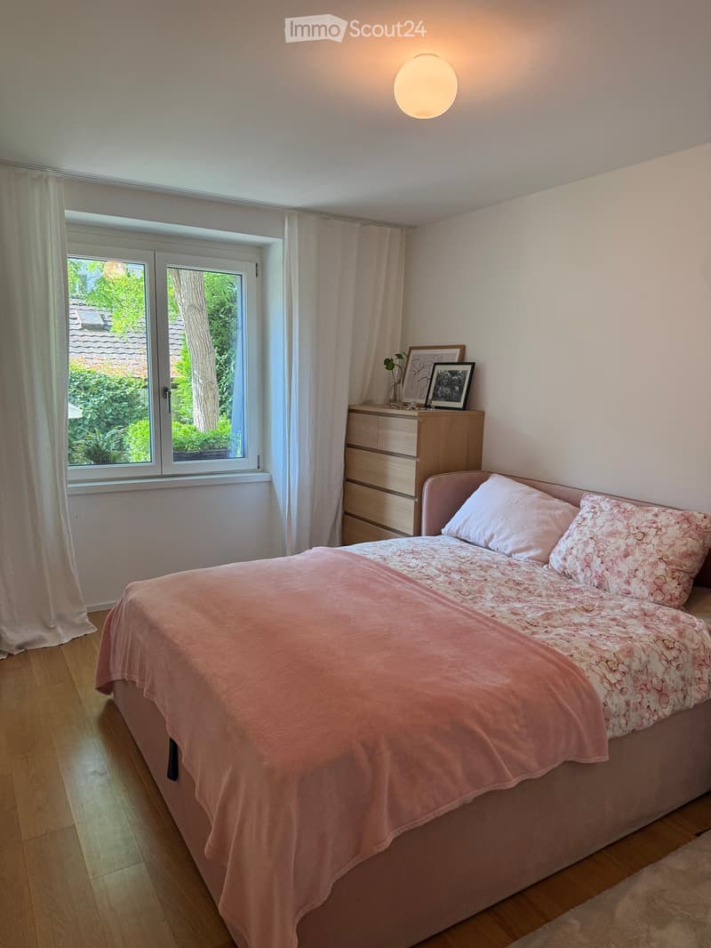 A well-lit bedroom with a double bed covered in a pink duvet and matching pillows, a wooden nightstand with a lamp, picture frames, and a vase with a small plant. The room features wooden flooring and large windows with white curtains.