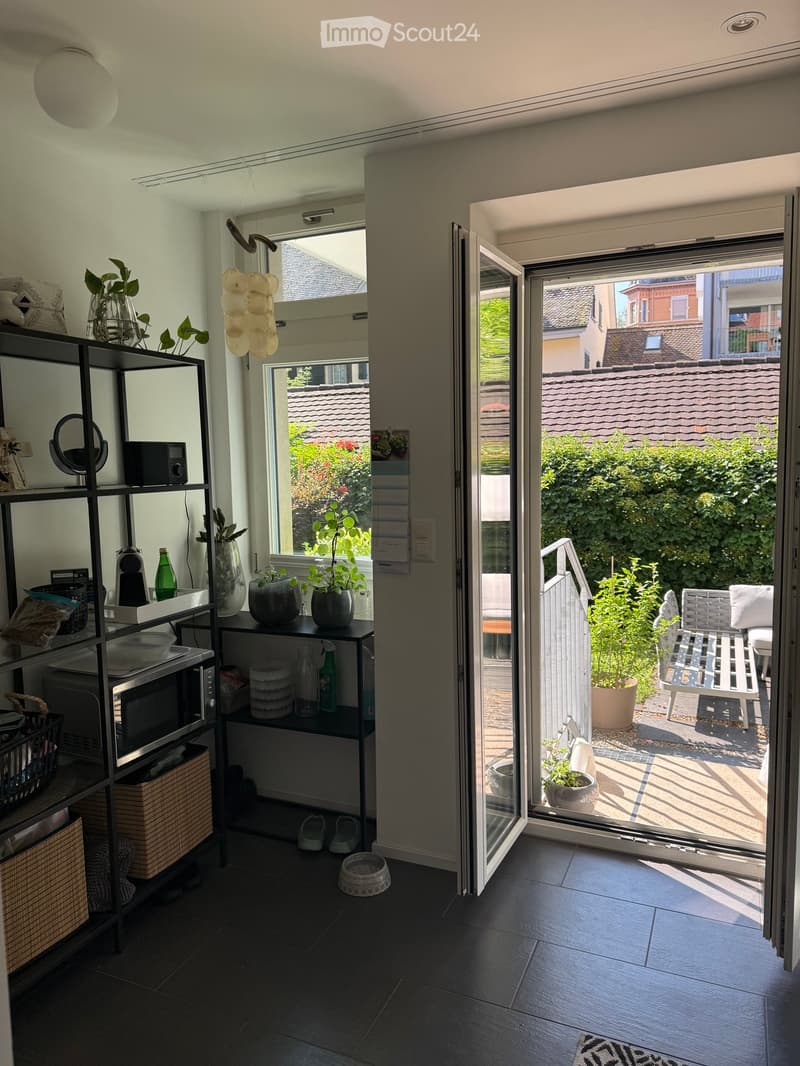 Modern interior with dark tiled floor, open sliding glass door leading to a balcony or terrace area with outdoor furniture and plants. Visible objects include a shelf with various items, a mirror, a radio, a coffee maker, a bottle, vases, potted plants, a