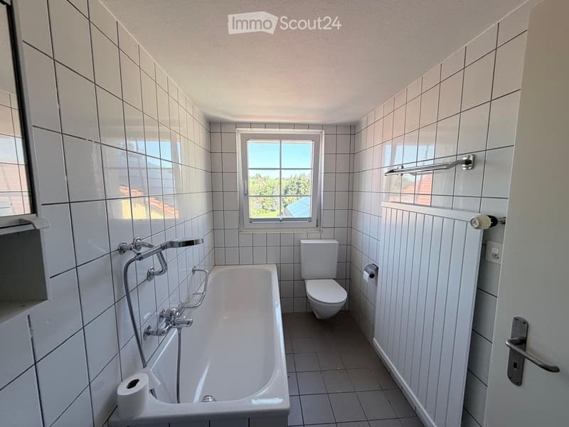 Bathroom with white tiled walls and floor, bathtub, toilet, and a window offering a view of greenery outside.