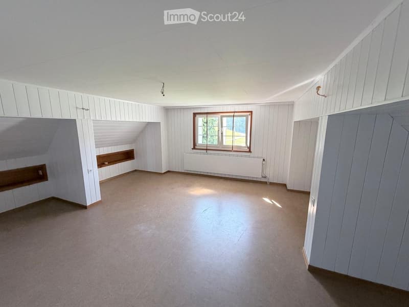 Empty room with white paneled walls, a window with wooden frame, and a radiator below it. The floor is concrete, and there is a small built-in shelf on one wall.
