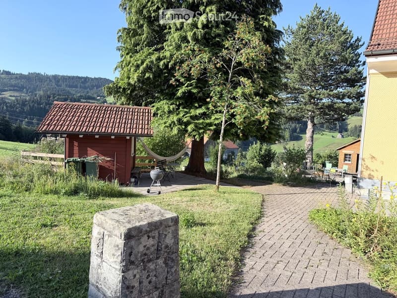 A peaceful garden area with a brick pathway, a hammock, a barbecue, and a stone pillar, surrounded by trees and a scenic countryside view.