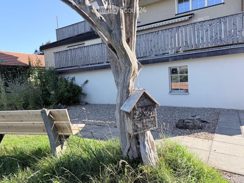 Garden area with a wooden bench, beehive, and a large tree trunk, near a two-story house with balconies and windows.