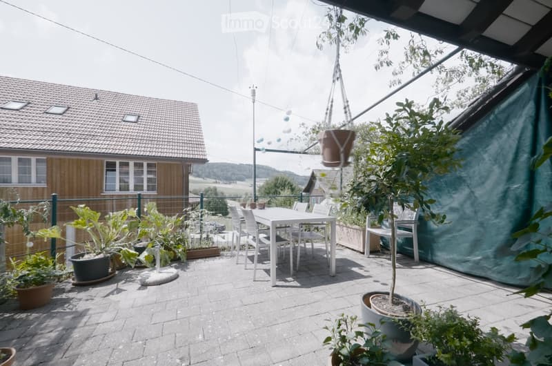 A covered outdoor patio with a white dining table and chairs, surrounded by potted plants and greenery, with a view of a house and countryside.