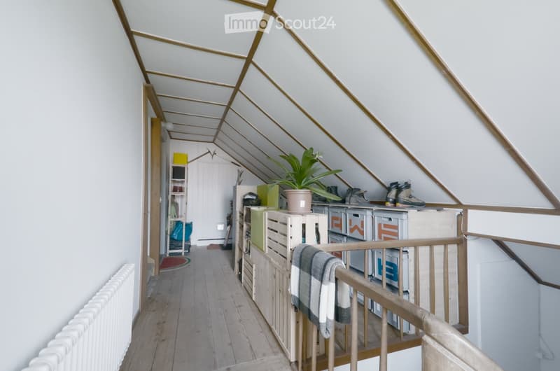 Attic corridor with wooden flooring, white walls, slanted ceiling with exposed wooden beams, storage crates, a potted plant, and a folded blanket on the railing.
