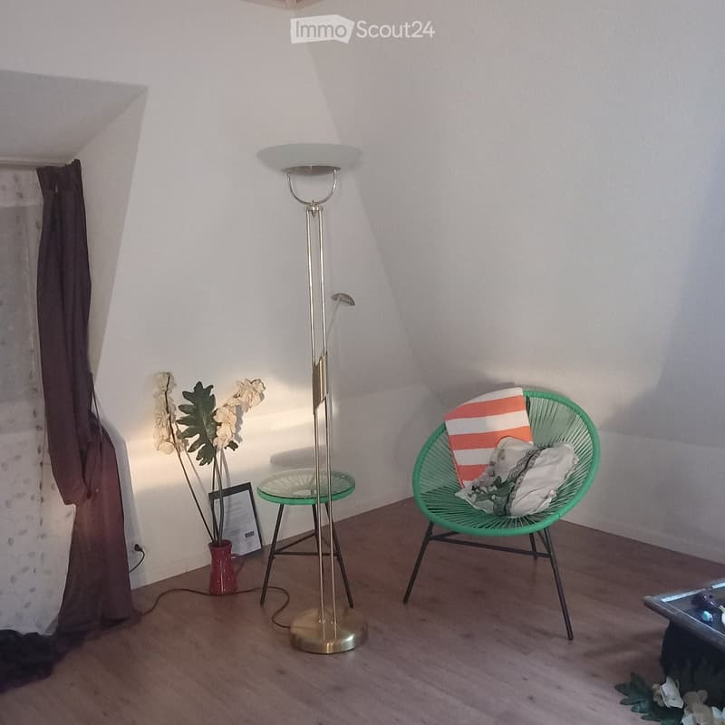 A small, cozy room with a green wicker chair, a glass side table, a floor lamp, a vase with flowers, and a picture frame on the floor.