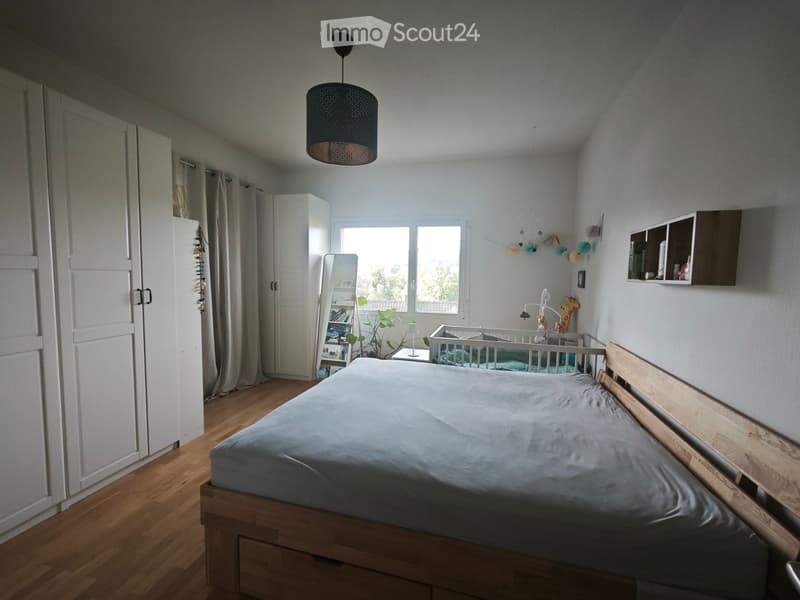 Cozy bedroom with wooden bed, wardrobe, mirror, small table with lamp, and baby crib with toys. Light-colored walls and wooden flooring.