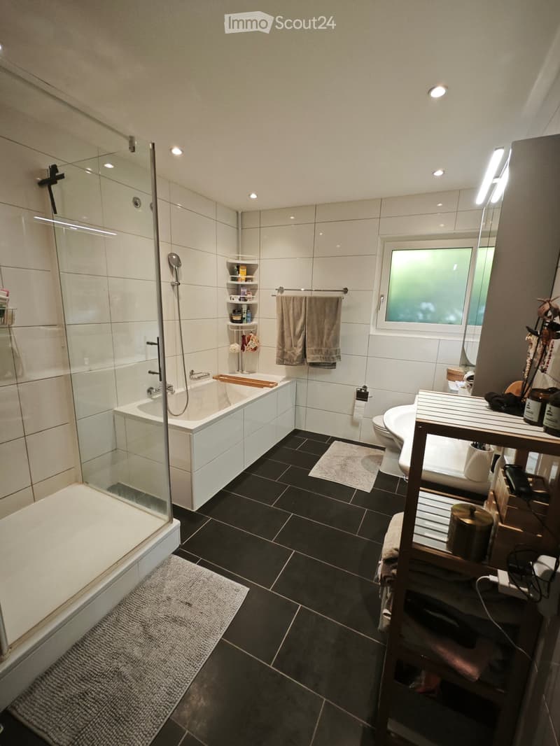 Modern bathroom with a bathtub, shower, toilet, sink, and a large mirror. Features include a wooden shelf, towels, and a small rug on the floor.