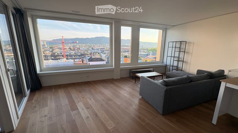 Modern living room with wooden flooring, large windows offering a city view, grey sofa, coffee table, and a tall shelf.