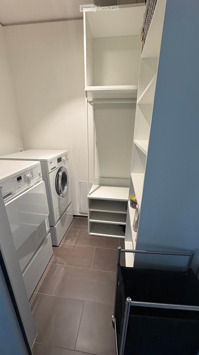 A small, well-lit room with a washing machine, dishwasher, and built-in shelves. The floor is tiled, and the walls are painted in neutral tones with one accent wall in blue.