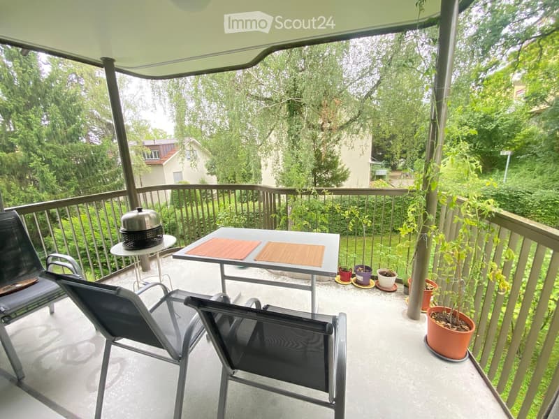Balcony with a table, chairs, and potted plants, featuring a covered area with a railing and greenery in the background.