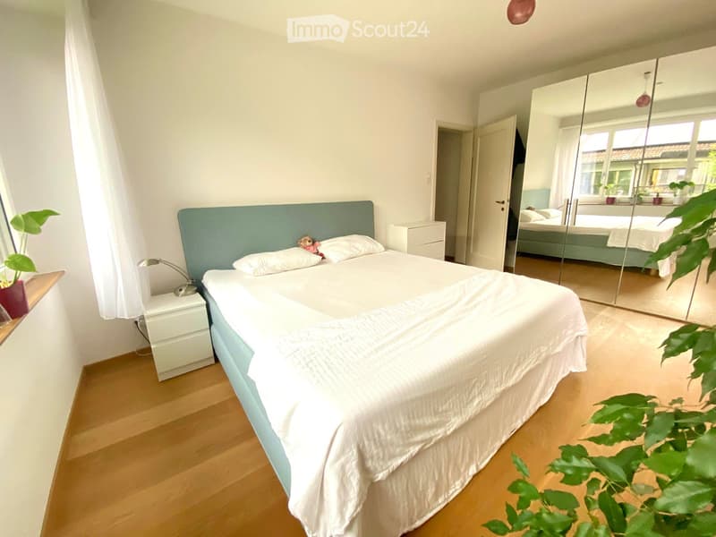 Bright bedroom with a double bed, white bedding, a nightstand with a lamp, a large mirror, and a potted plant near the window.