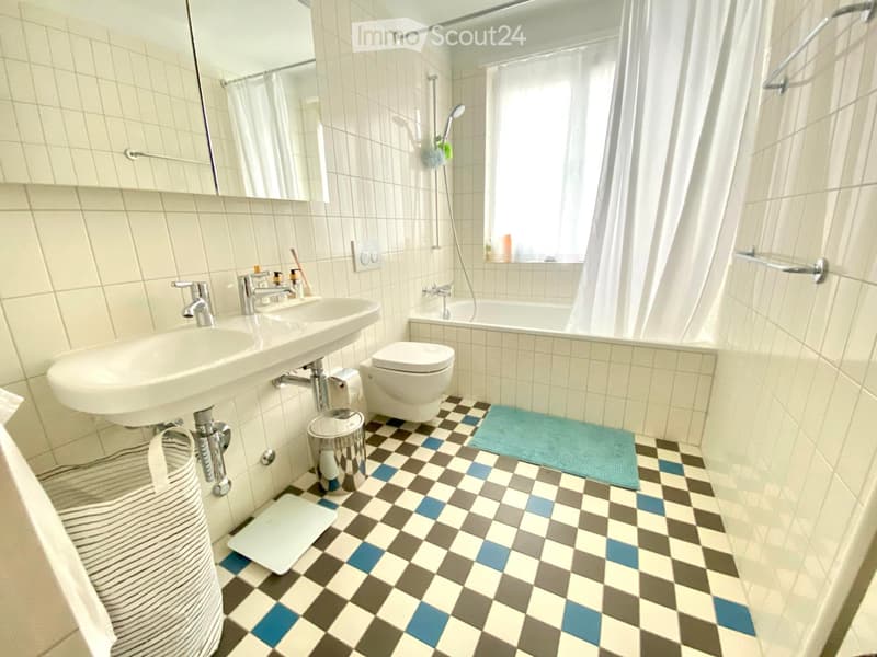 Bathroom with a sink, toilet, bathtub, and shower area. Features a checkered floor with blue, gray, and white tiles, a white tiled wall, and a mirror above the sink.