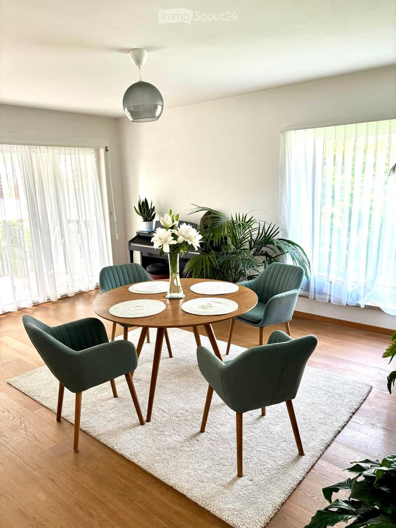 Dining room with a round wooden table, four green chairs, a white rug, and a vase with white flowers on the table. Large windows with white curtains and a hanging lamp are visible.