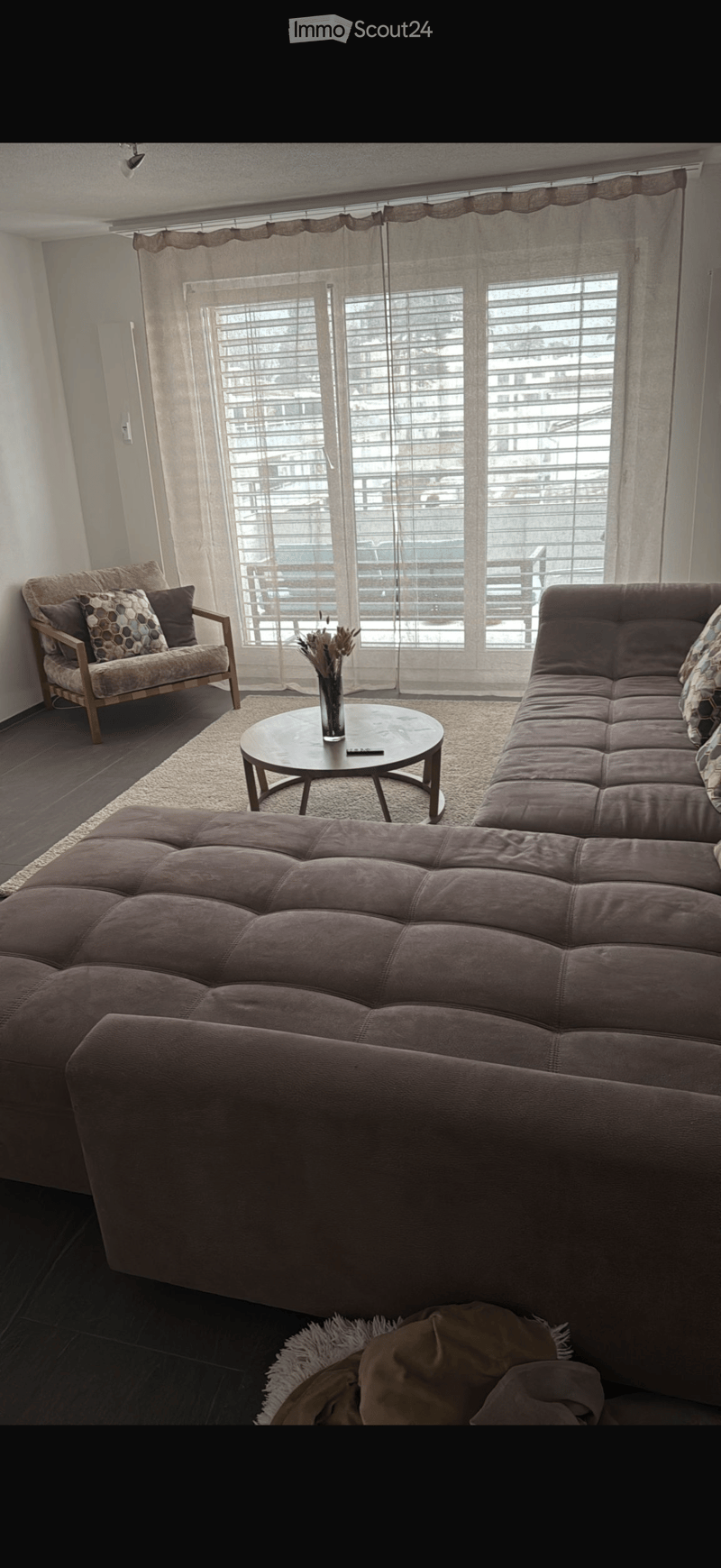 Modern living room with large windows, a sectional sofa, a wooden armchair, a round coffee table with a vase, and a blanket on the floor.