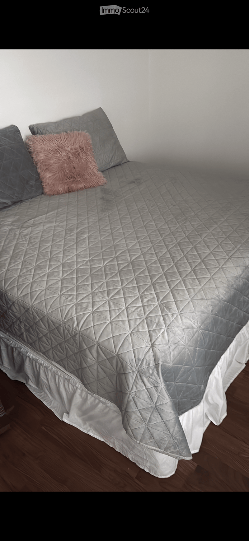 A neatly made bed with a grey quilted comforter, two grey pillows, and a pink throw pillow on a wooden floor.