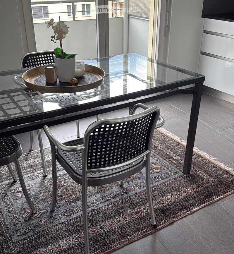 Modern dining area with a glass table, metal chairs, a decorative tray with a flower pot and candle, and a patterned rug.