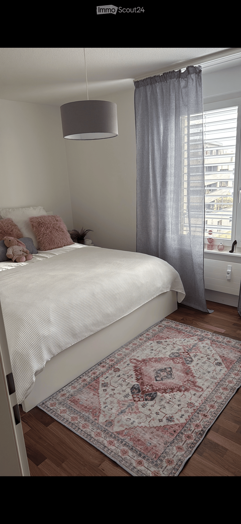 Cozy bedroom with a large bed, patterned rug, soft pillows, and a hanging lamp. The room features wooden flooring and large windows with grey curtains.