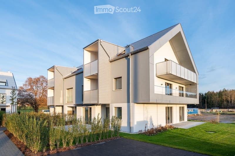 Modern multi-story residential building with clean lines, large windows, and balconies. The structure features a combination of light grey and white finishes, with a landscaped front yard and paved walkway.