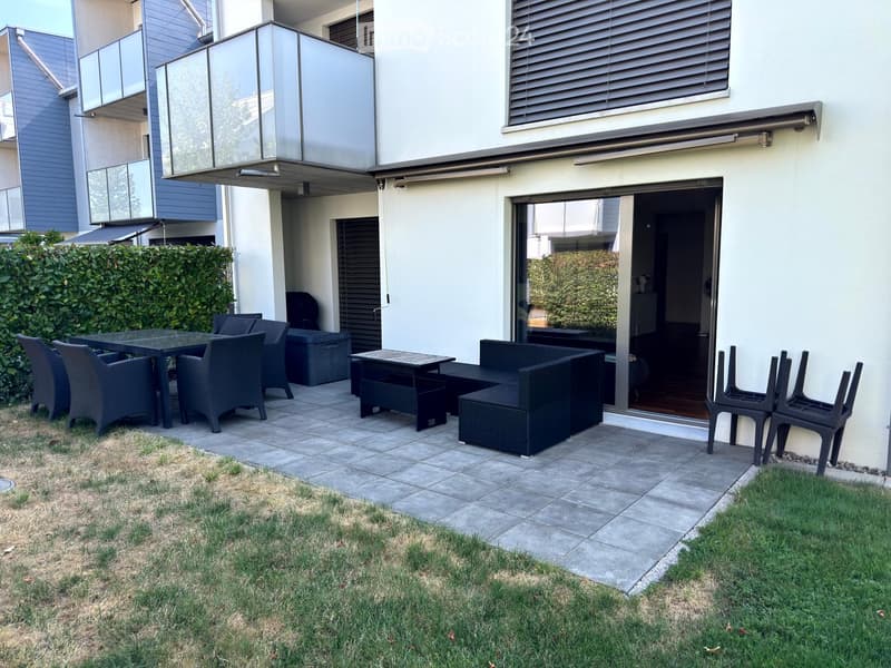 Outdoor patio area with a dining table, chairs, sofa, and stools, adjacent to a modern building with large windows and a glass balcony.