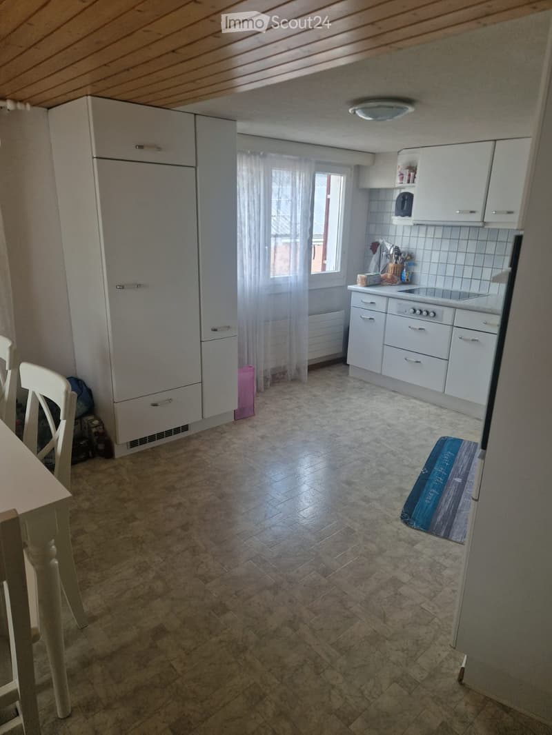 Modern kitchen with white cabinets, a refrigerator, an oven, a sink, and a dining area with a table and chairs. The floor is tiled, and there is a window with white curtains.