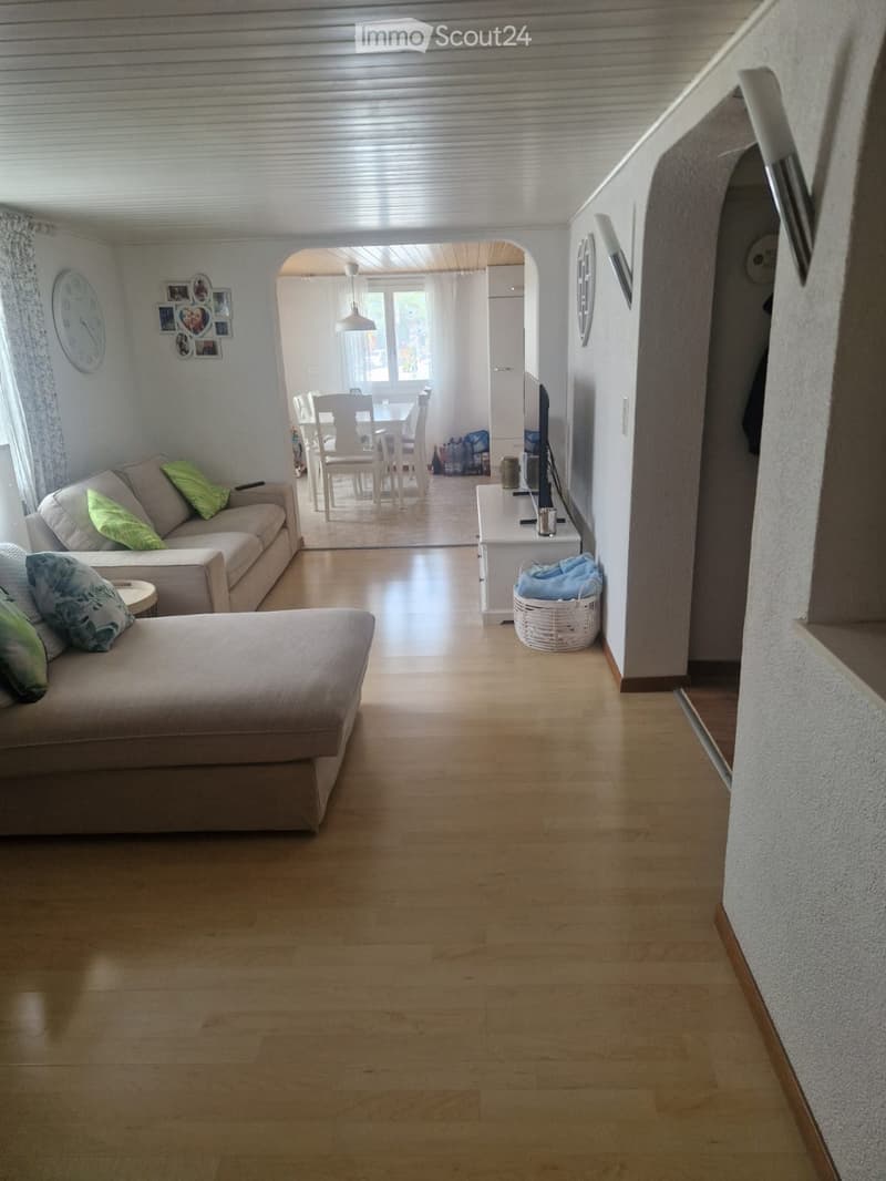 Bright living room with wooden flooring, a sofa, ottoman, coffee table, dining area with a table and chairs, and a television on a stand. A white basket is placed near the television stand.