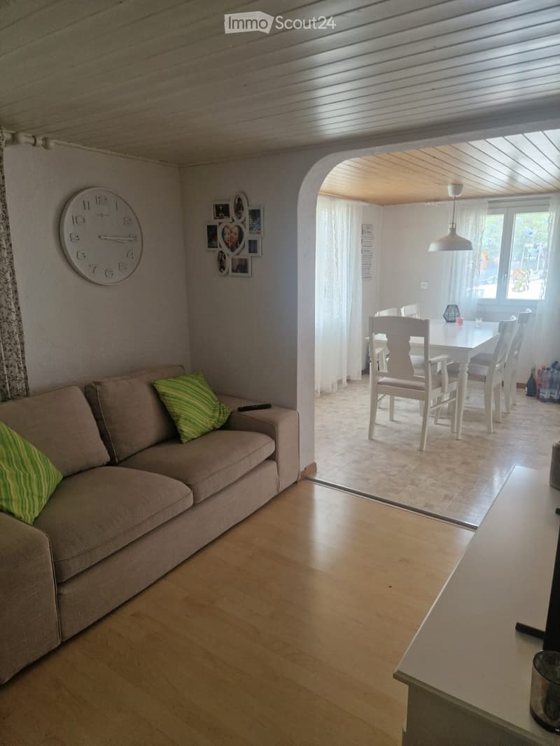 Living room with a sofa, green cushions, a clock on the wall, and a dining area with a table and chairs visible through an archway.