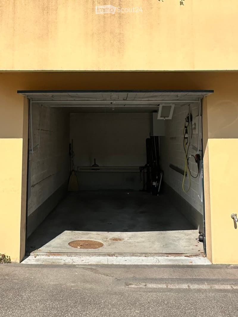 Open garage with concrete floor, light-colored walls, and some stored items including a vacuum cleaner and a small table.