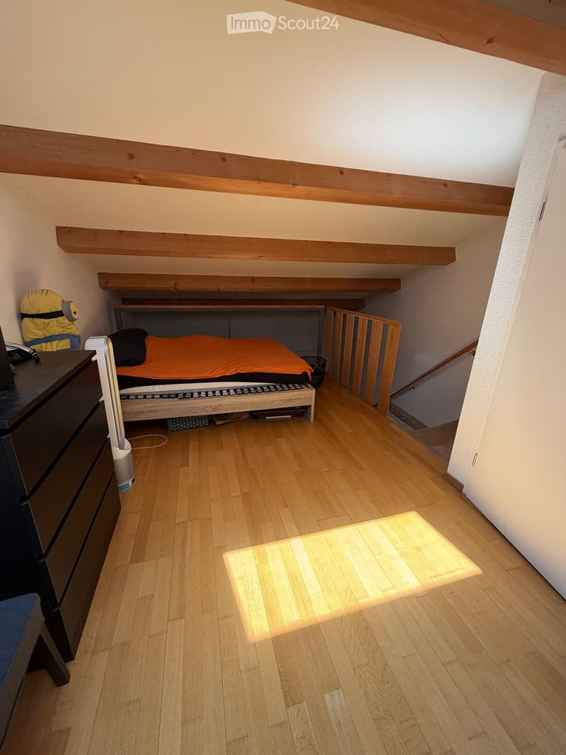 Cozy attic bedroom with wooden floors, slanted wooden ceiling beams, a single bed with orange bedding, a nightstand with a lamp, and a wardrobe. A small staircase leads to another level.