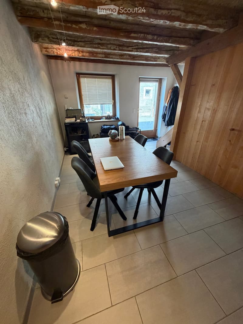 Dining area with a wooden table, four chairs, a laptop, a small cabinet, and a trash can. The room has exposed wooden beams on the ceiling and a window with blinds.
