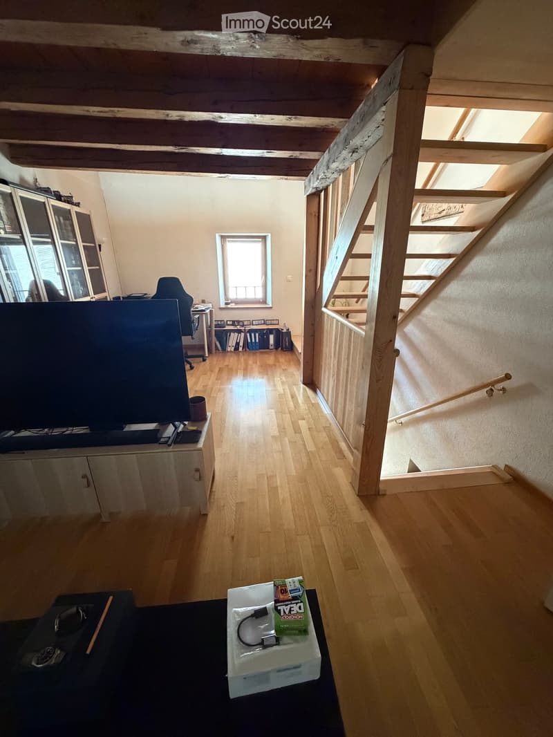 Bright living room with wooden floors, exposed wooden beams, TV on stand, chair, small table, and cabinet. Stairs lead to another level.