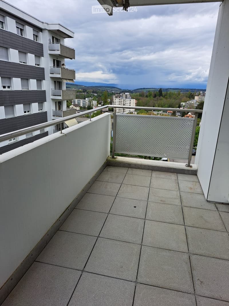 Balcony with tiled floor, metal railing, and a view of a multi-story residential building and surrounding landscape.