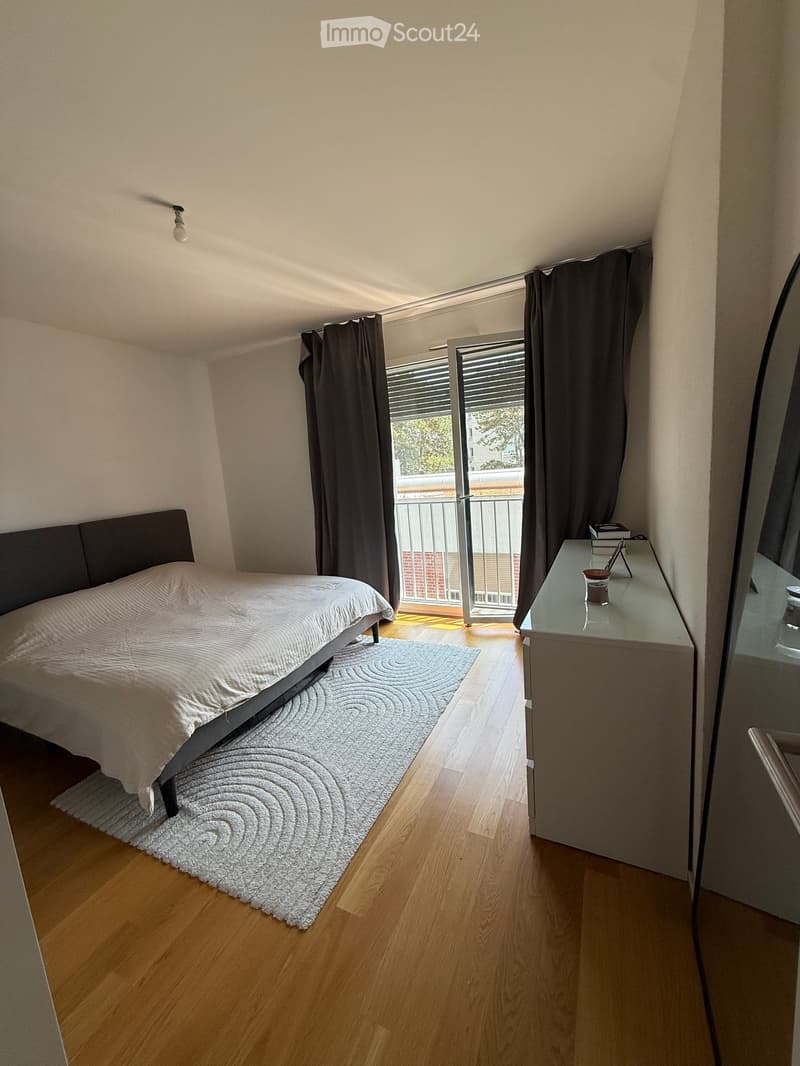 Modern bedroom with wooden flooring, a bed with a dark headboard, a nightstand with a mirror, and a balcony with dark curtains.