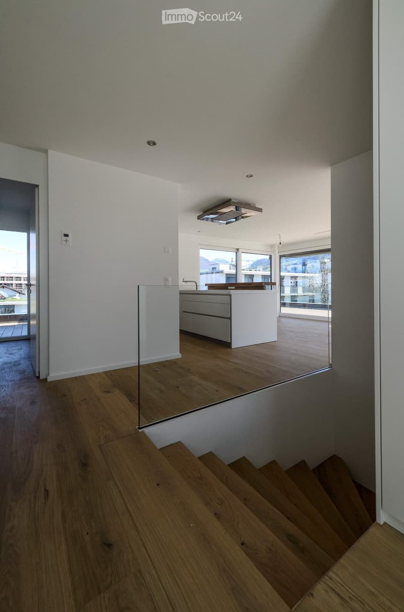 Modern kitchen with a white island, wooden flooring, and large windows overlooking a balcony. The space features a sleek, minimalist design with a glass partition separating the kitchen from the living area.