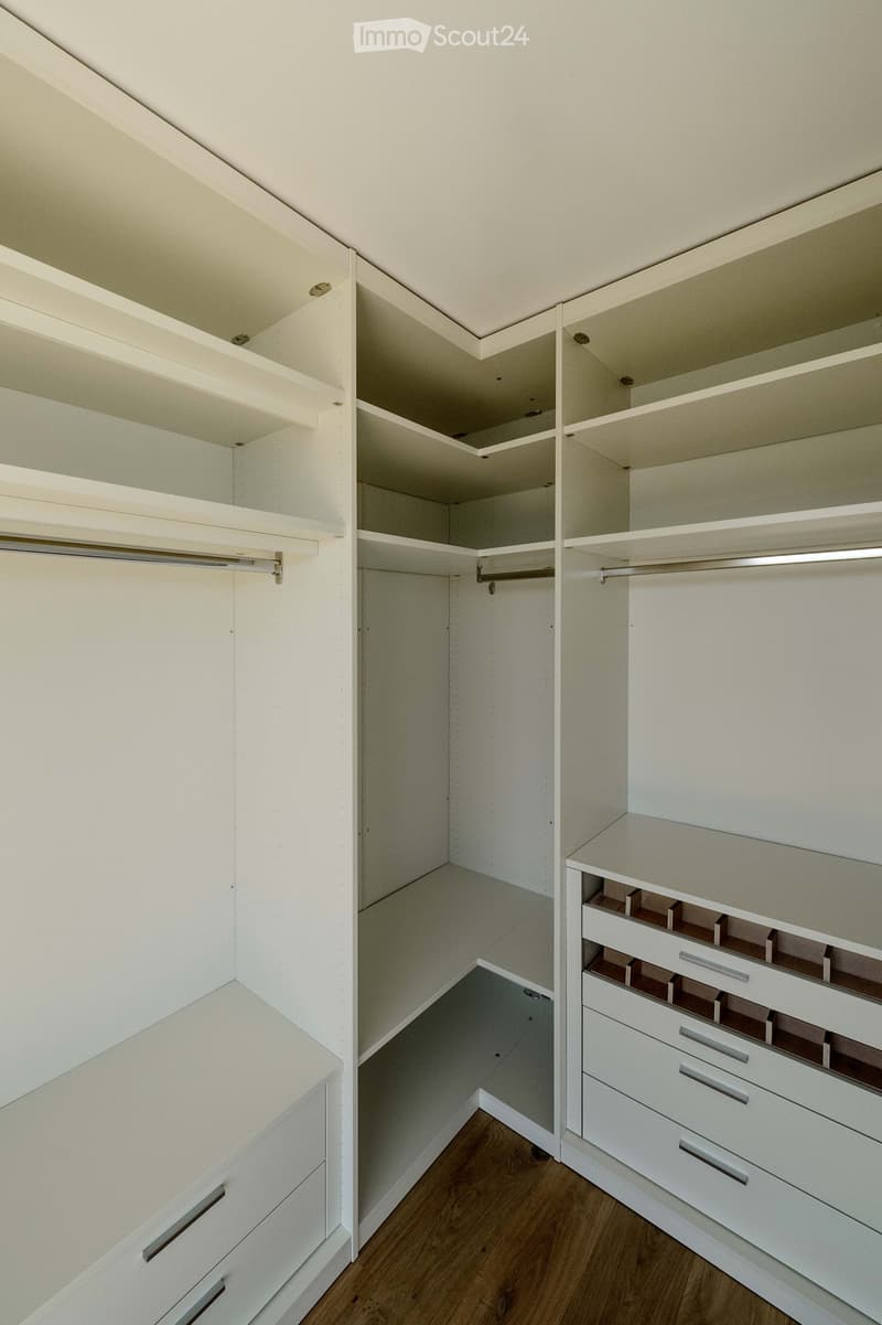 Empty walk-in closet with white built-in shelving units, drawers, and hanging rods. The space features a wooden floor and neutral-colored walls.