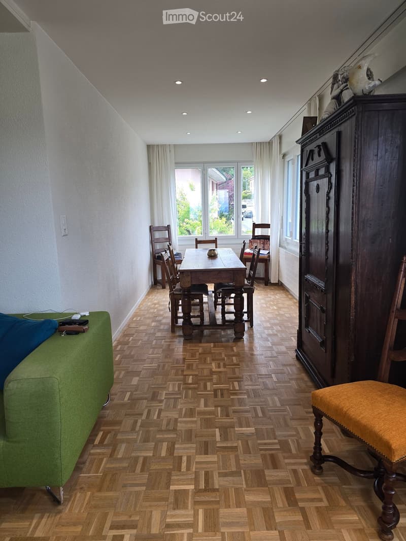 A well-lit room with wooden herringbone flooring, a green sofa, a wooden dining table with chairs, and a tall dark wooden cabinet.