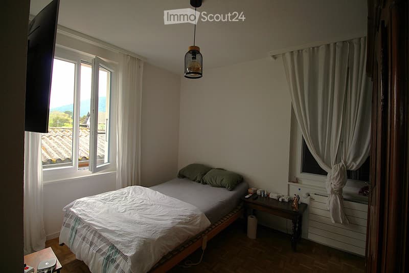 Cozy bedroom with a single bed, nightstand, small table, and large windows with curtains. The room features wooden flooring and a view of the roof and landscape outside.