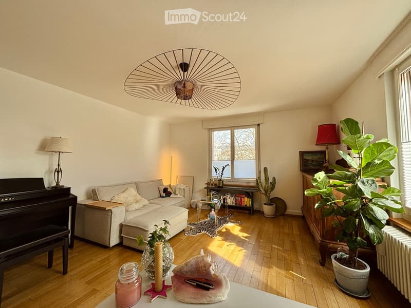 Bright living room with wooden flooring, large window, grey sofa, piano, decorative plants, and a modern hanging light fixture.