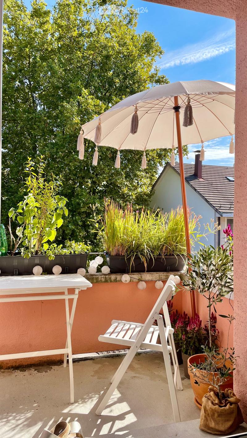 A sunny balcony with a white table, white folding chair, large umbrella, and various potted plants.