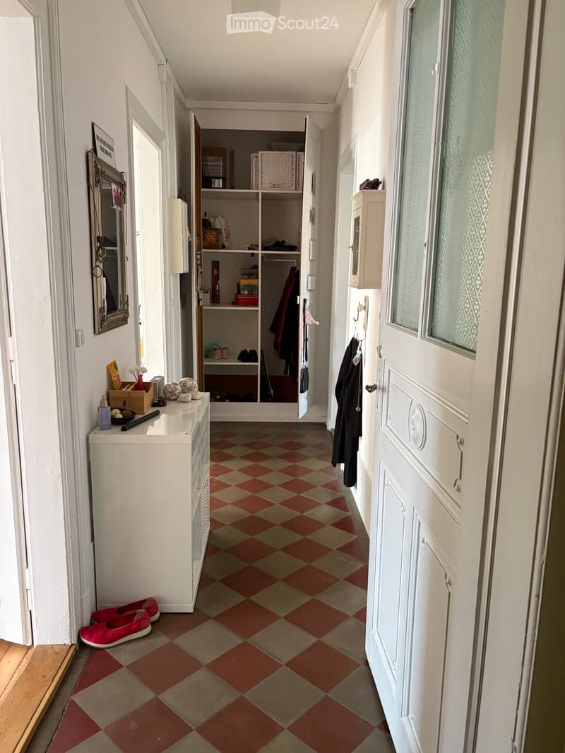 Hallway with checkered tile floor, white walls, open closet with clothes and storage items, small table with decorative items, and a coat rack with jackets.