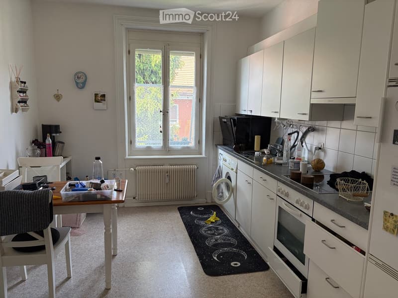The kitchen features white cabinets, a black countertop, and various appliances including an oven, fridge, and dishwasher. A small wooden table with a chair is positioned near the window, and a black rug with a moon and stars design lies on the floor.