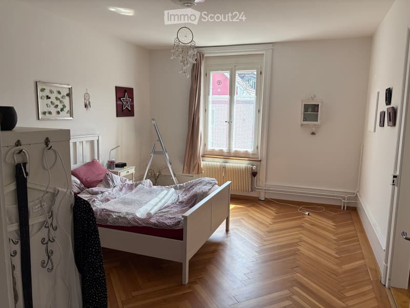 Cozy bedroom with wooden flooring, a white bed with grey bedding, a nightstand with a lamp, a radiator under a window, and decorative items on the walls.