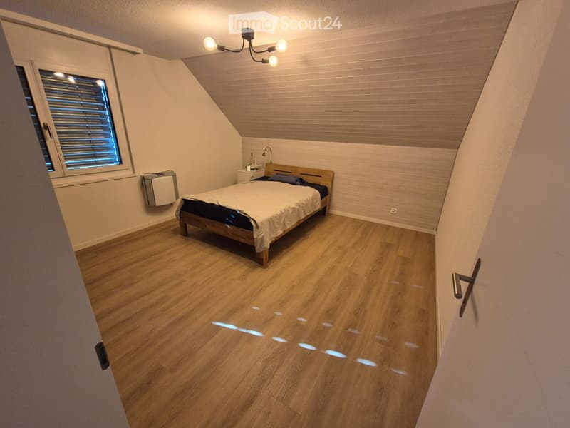 Cozy attic bedroom with wooden floors, slanted wooden ceiling, large window with blinds, bed with white bedding, nightstand, and wall-mounted lamp.
