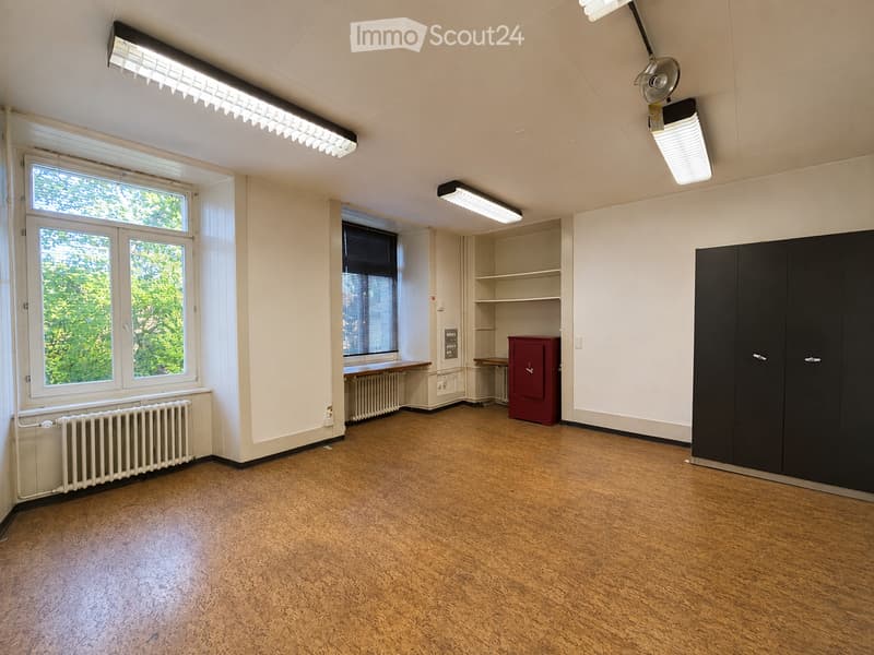 Empty room with wooden flooring, white walls, large windows, built-in shelving, and a small red cabinet.